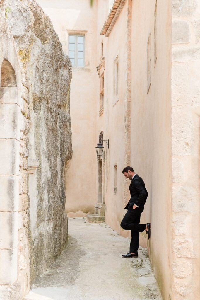 Portrait décontracté et amusant du marié en smoking noir dans une ruelle de village provençal, pose décontractée contre un mur en pierre de taille