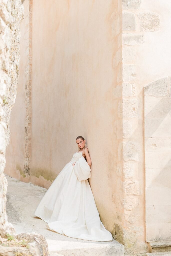 Portrait minimaliste de la mariée contre un mur ancien de Provence, robe volumineuse en soie blanche aux lignes épurées, pose élégante et sophistiquée