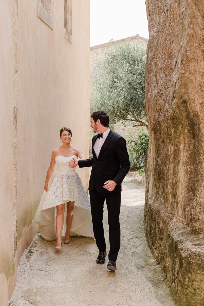 Jeunes mariés marchant main dans la main dans une ruelle pittoresque de Provence, entre des murs de pierre ocre et des oliviers, la mariée portant une robe courte devant asymétrique avec détails en dentelle