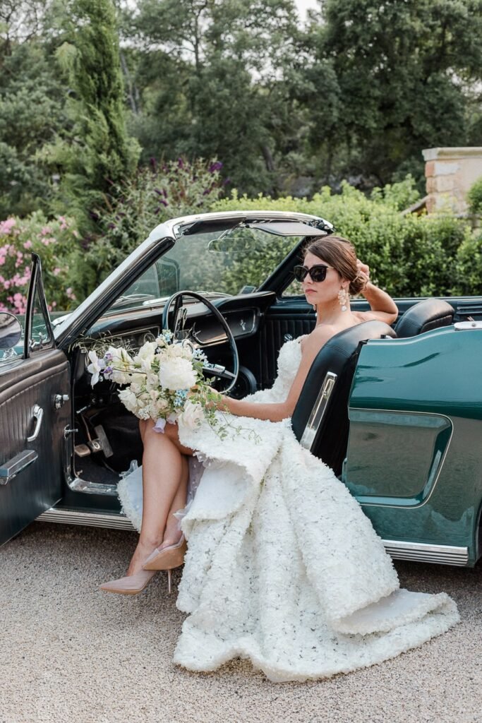 Mariée élégante en robe blanche texturée assise dans un cabriolet vintage vert foncé, portant des lunettes de soleil et tenant un bouquet blanc, entourée de la verdure méditerranéenne provençale