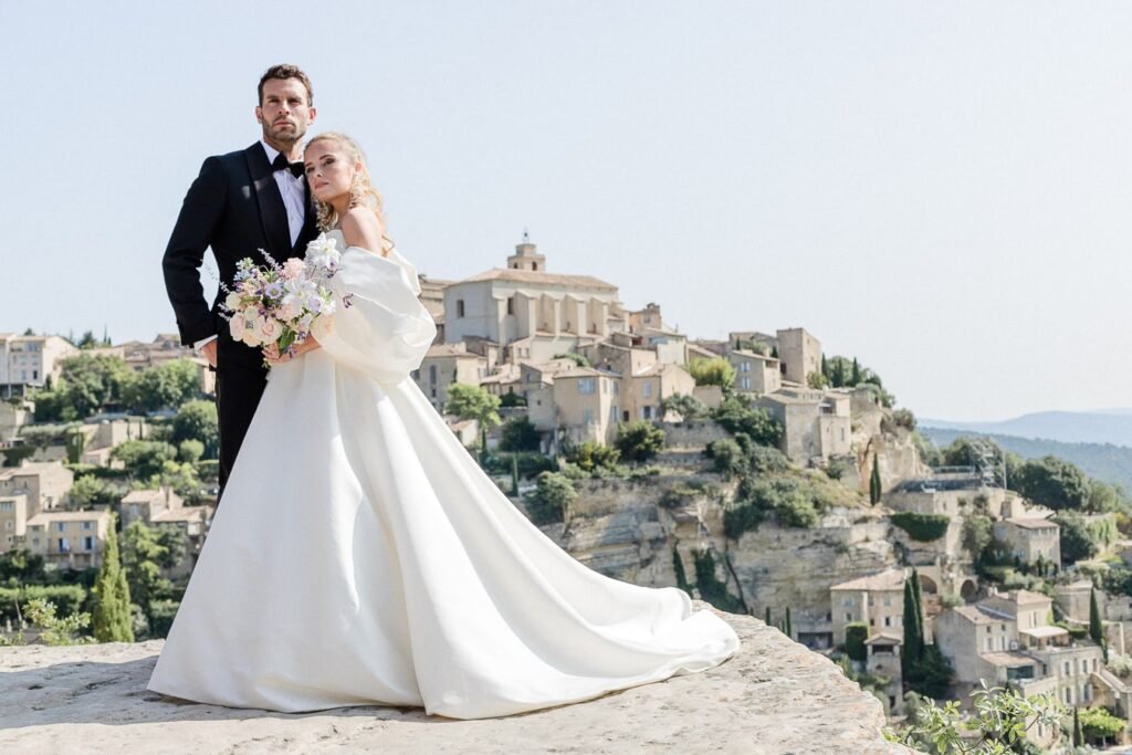 Couple de mariés élégants surplombant le village perché de Gordes, elle en robe blanche architecturale avec bouquet pastel, lui en smoking noir, vue panoramique sur le Luberon