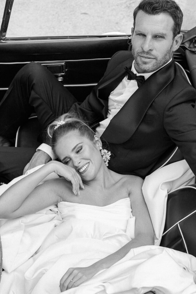 Portrait glamour en noir et blanc des mariés dans une voiture de collection, lui en smoking et elle en robe structurée, sourire complice et naturel