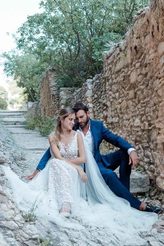Jeunes mariés complices sur des marches en pierre ancienne, elle en robe délicate de dentelle blanche, lui en costume bleu, cadre naturel et authentique de Provence