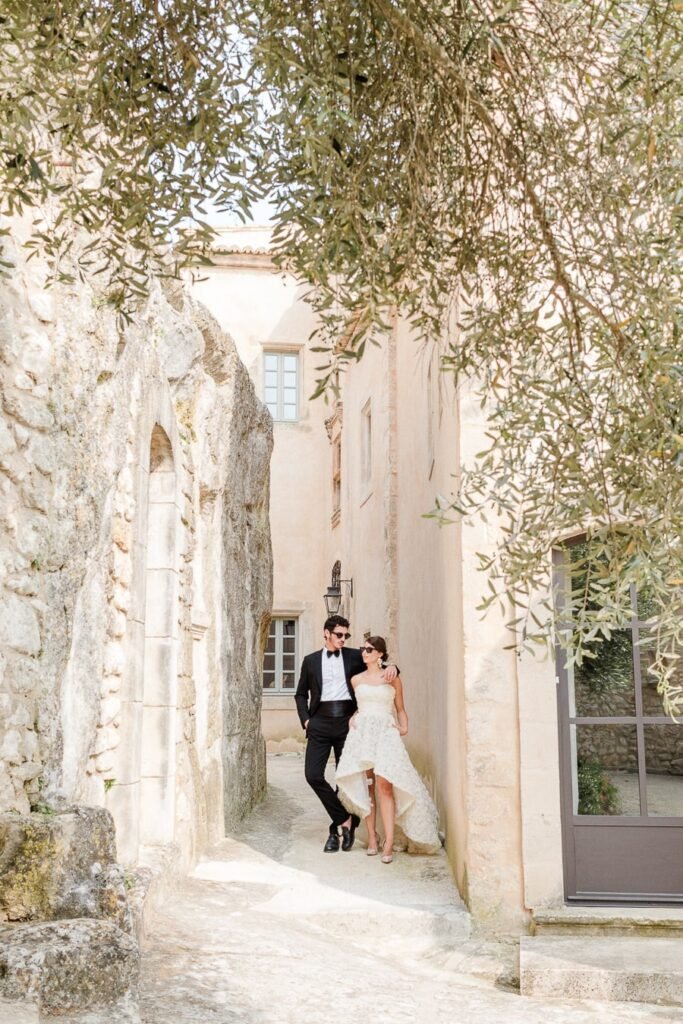 Couple de mariés élégants dans une ruelle de château provençal aux murs beige, encadrés par des branches d'oliviers, la mariée en robe courte asymétrique et le marié en smoking noir