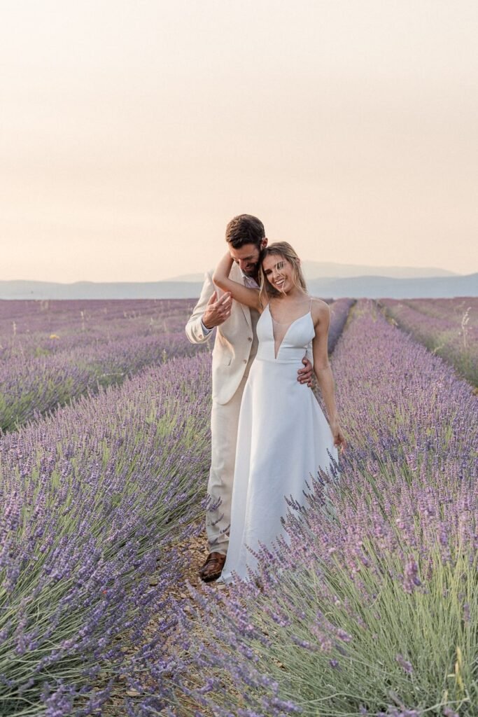 Jeunes mariés complices photographiés au coucher du soleil dans un champ de lavande de Valensole, elle en robe blanche épurée, lui en costume beige clair. Un souvenir précieux capturé par un photographe de mariage proposant des tarif adaptés