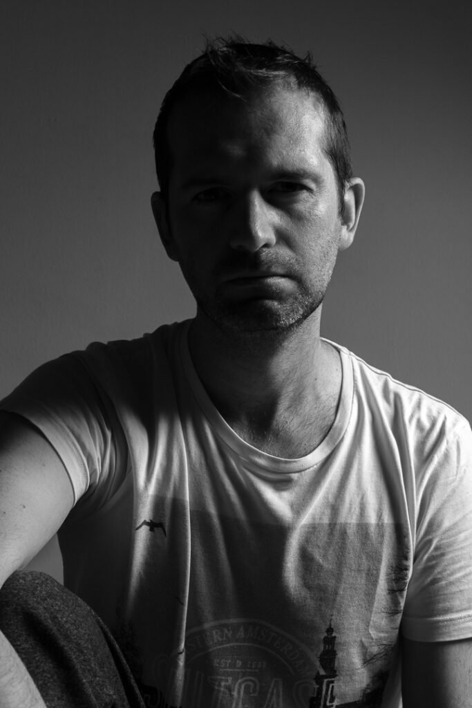 Portrait noir et blanc contrasté d'un homme en t-shirt blanc, photographe de mariage en Provence, avec un éclairage latéral dramatique mettant en valeur ses traits et son expression déterminée