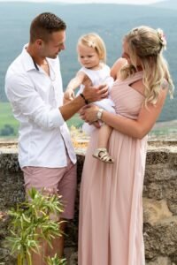 Jeune famille élégante posant sur une terrasse en pierre ancienne, parents et enfant coordonnés en tons blancs et roses, vue sur les collines provençales