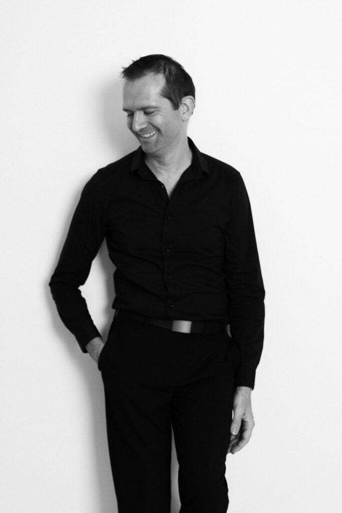 Portrait noir et blanc d’un photographe de mariage, souriant et regard baissé, vêtu d’une chemise noire, posé contre un mur blanc.