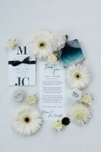 Papeterie de mariage élégante entourée de fleurs blanches, alliances, parfum et message de remerciement personnalisé.